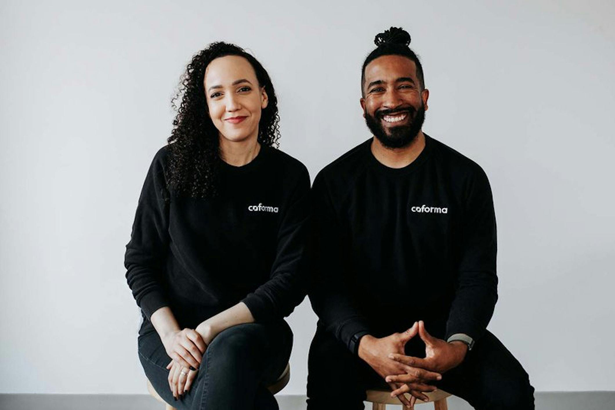 Ashleigh Axios and Eduardo Ortiz sit warmly smiling at the viewer in matching sweaters with the Coforma logo on the chest